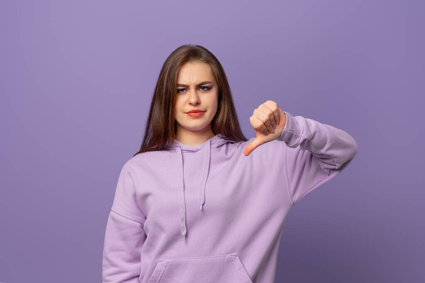 Huonoja uutisia. Tyytymätön nuori brunette nainen osoittaa peukalonsa alas paheksuen tarjouksen tilannetta, kun seisoo violetti tausta. Kielteiset ihmisen tunteet, kasvojen tunteet - Valokuva, kuva