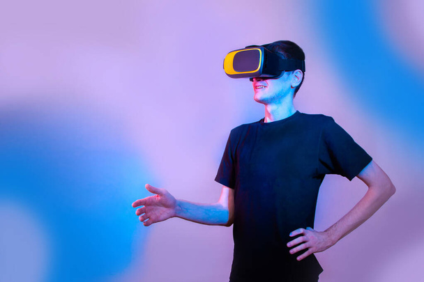 A young man in virtual reality glasses gives a hand to say hello. Communication in virtual reality. Neon colors, copy space - Foto, immagini