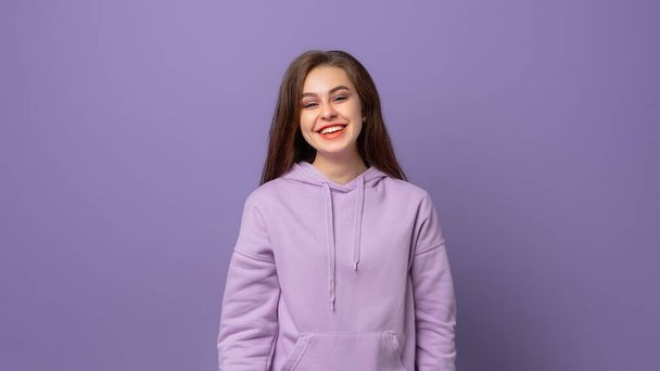 Portrét mladé ženy při pohledu na kameru s čistou pletí, přirozeným make-upem a velkým úsměvem, stojící nad fialovým pozadím - Fotografie, Obrázek