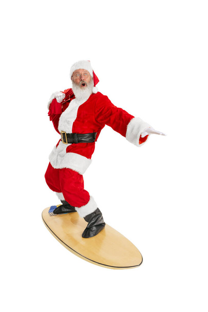 Portrait of emotional Santa Claus in red-white costume standing surfboard isolated on white background. Merry Christmas concept - Foto, Imagen