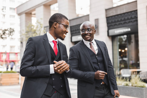 Two dark-skinned african american businessmen in suits and glasses walk outdoors in the city and discuss business - Photo, Image