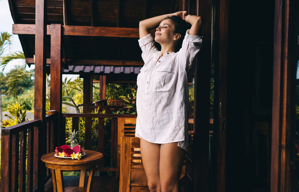 Satisfied young female with closed eyes in white shirt enjoying nature and breathing fresh air while standing on terrace of wooden cottage with hands over head - Фото, зображення