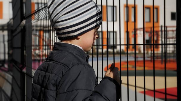 Upset lonely boy looking through metal fence on children palyground and modern school - Photo, Image
