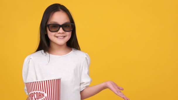 Счастливая маленькая азиатка в 3D очках с ведром попкорна показывает что-то с протянутой рукой, свободное место - Кадры, видео
