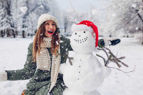 Happy woman toss snow up by snowman in Santa hat outdoors in snowy winter park. Christmas festive season. Fun activities - Photo, Image
