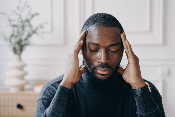 Uitgeput jonge Afro Amerikaanse mannelijke ondernemer zitten met de ogen dicht en met de handen op zijn hoofd - Foto, afbeelding