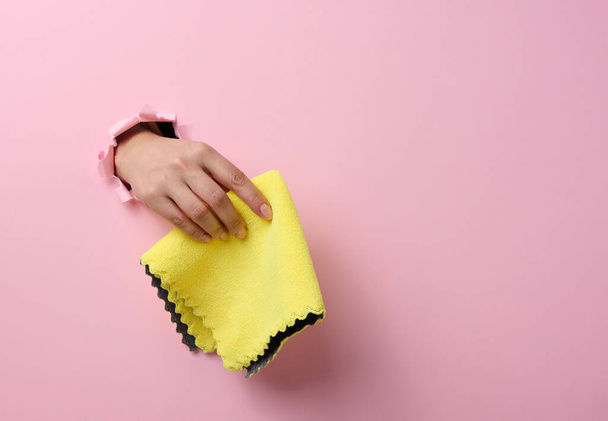 mão feminina saindo de um buraco rasgado em um fundo de papel rosa e segurando um pano seco - Foto, Imagem