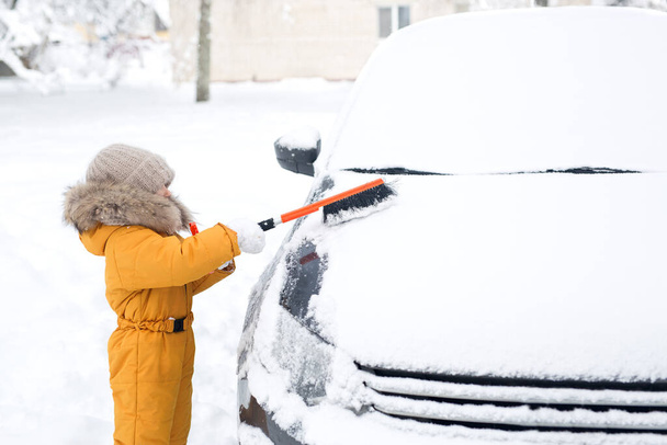 Dziecko w pomarańczowej kurtce z futerkiem i czapce z dzianiny w rękawiczkach czyści śnieg z samochodu przed wyjazdem. Czyszczenie pojazdu pokrytego śniegiem i lodem szczotką do czyszczenia - Zdjęcie, obraz