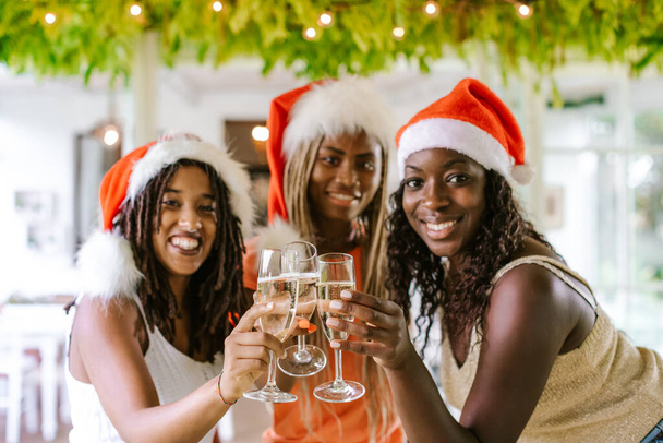 Happy smiling African American girls celebrating Christmas (or new year) and cheering with champagne. Winter holidays and multiracial group concept. Focus on glasses - Photo, image