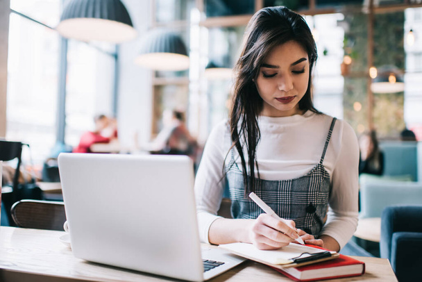 Focused female freelancer in casual clothes sitting at table in light cafe and taking notes on clipboard while working on project - Foto, imagen