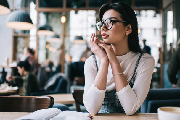 Young serious female student in casual outfit with eyeglasses looking away while sitting at table in cafeteria leaning on hands - Фото, изображение