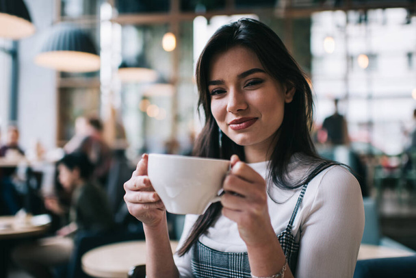 Happy female in casual clothes looking at camera while sitting with mug of coffee on blurred background of modern cafeteria - 写真・画像