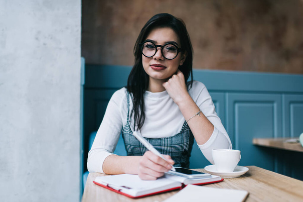 Content woman in casual wear and eyeglasses looking at camera and writing in notebook while working remotely with smartphone in cafe - Foto, immagini