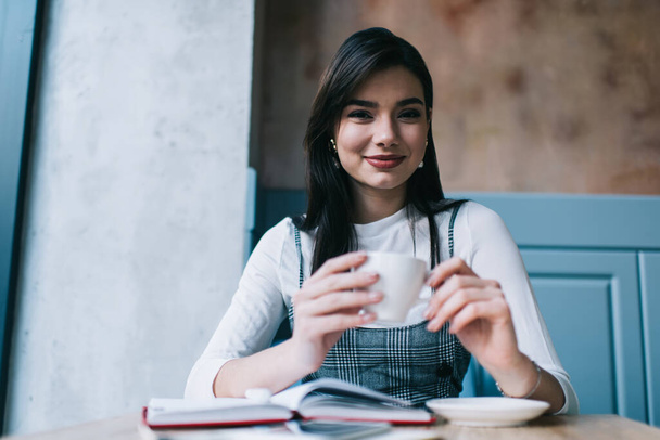 Smiling young woman with dark hair in stylish clothes drinking cup of tea and looking at camera while sitting at table with opened notebook in cafe in daylight - Fotografie, Obrázek