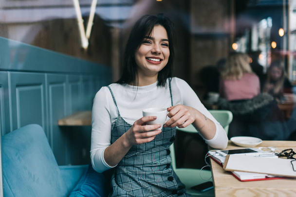 Happy young female student wearing trendy clothes sitting at table with notebook and papers and drinking hot beverage while preparing for exams in cafe - Foto, imagen