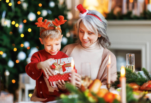 Großmutter mit aufgeregtem Enkelkind beim Auspacken der Weihnachtsgeschenkbox neben geschmücktem Weihnachtsbaum - Foto, Bild