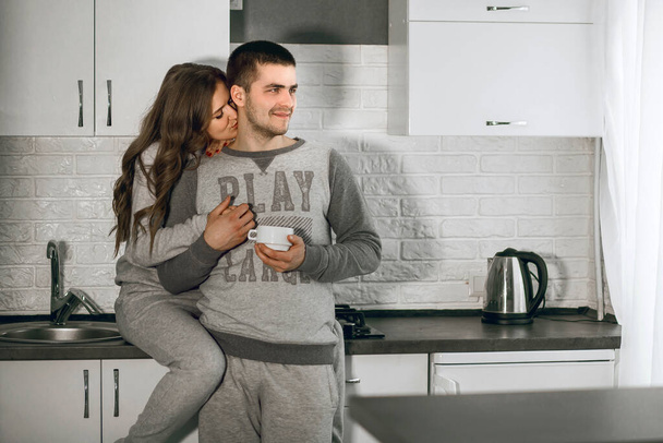 Молодая влюбленная пара завтракает на кухне и обнимается. Мужчина пьёт кофе, а девушка целует его в шею. Любовные отношения гетеросексуальной пары. Счастливая супружеская пара - Фото, изображение