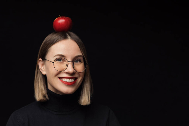 Donna sorridente che guarda alla fotocamera con una mela sulla testa, indossa occhiali su sfondo nero - Foto, immagini