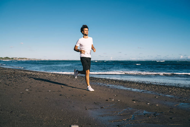 Full length young sportsman in sportive clothes training and running alone on pebble beach near waving sea on sunny cloudless day - Photo, image
