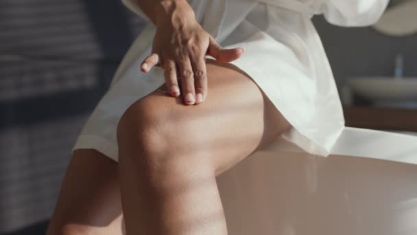 Péče o tělo. Mladá nerozpoznatelná černoška nanáší na nohy krém proti celulitidě, sedí v koupelně - Záběry, video