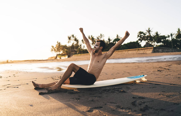 Excited surfer guy sitting at professional surfboard and raising hands in air satisfied with sportive goals during summer vacations in Miami, joyful man celebrating triumph during daytime at seashore - Foto, Imagen