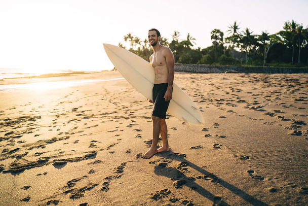 Full length of cheerful man with professional surfboard smiling at camera during weekend time for hobby practice, happy male surfer enjoying summer vacations at Malibu Beach in sunny California - Fotó, kép