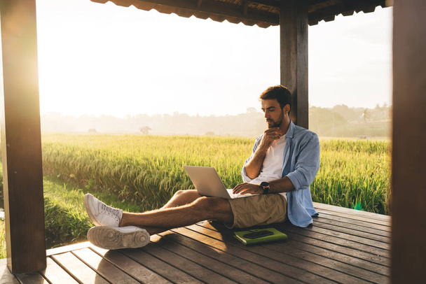 Pondering male remote employee surfing internet on portable computer while touching chin and sitting with crossed legs on wooden porch near bright grass field in sunlight - Φωτογραφία, εικόνα