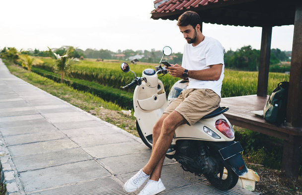 Young attentive male traveler text messaging on cellphone while sitting with crossed legs on vehicle on walkway near pavilion in summer - Foto, immagini
