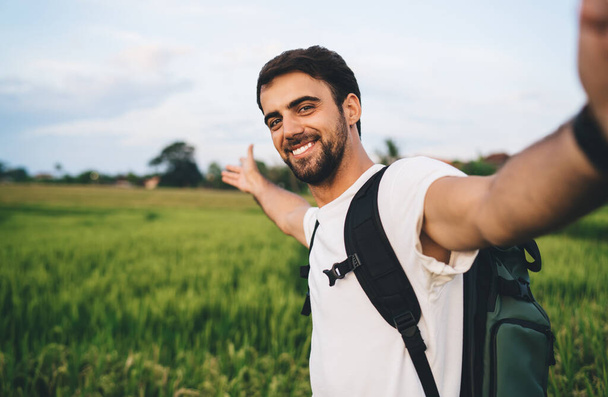 Side view handsome young male in casual white shirt with backpack taking selfie and showing green meadow while holding camera and looking with smile - Valokuva, kuva