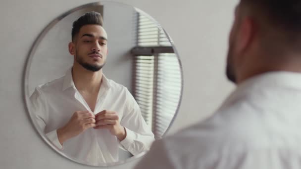 朝のルーティン。若い自信を持って中東のビジネスマンが鏡に笑顔で彼のシャツをボタンアップ - 映像、動画