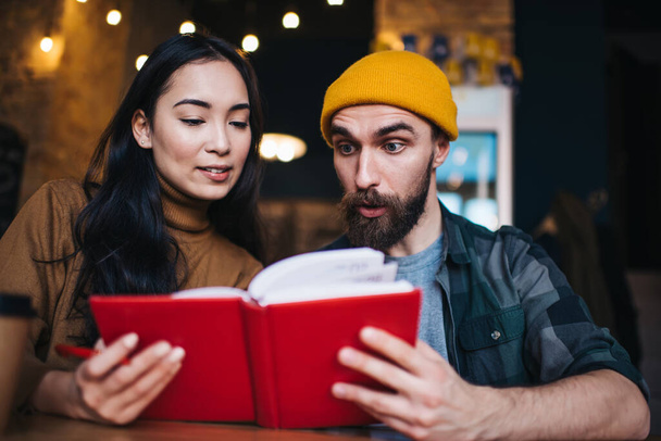 Amazed man in trendy cap shocked with education information from textbook, Caucasian male and female students 20 years old reading knowledge literature while preparing to college exams togetherness - Foto, Imagen