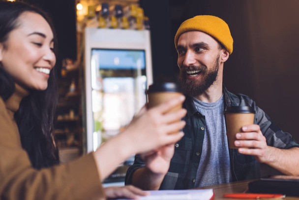 Positive bearded man in trendy hat and casual clothes sitting at wooden table in cafe and looking at girlfriend while drinking coffee from paper cups - Photo, Image