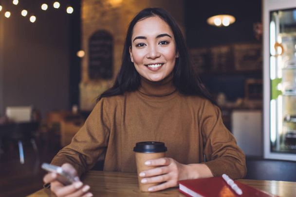 Asian female in casual outfit smiling at camera while drinking coffee and using smartphone sitting at table in cozy cafeteria - Fotografie, Obrázek