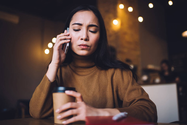 Serious female freelancer speaking on smartphone and holding cup of hot coffee during coffee break in cozy restaurant in evening time - Photo, Image