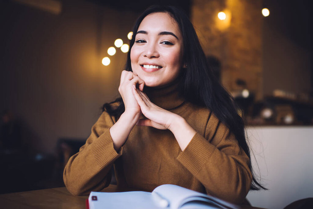 Happy youthful female in brown turtleneck sweater sitting in cafe with notebook on table while smiling and looking at camera - Фото, изображение