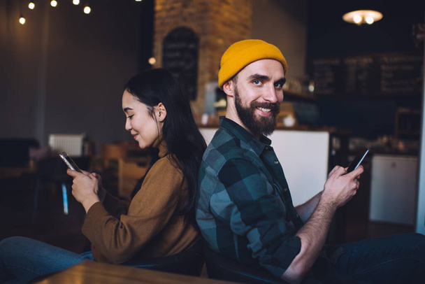 Portrait of cheerful male user with smartphone device in hand smiling at camera while female friend browsing booking website on mobile, millennial hipster guys connecting to 4g for reading content - Foto, Bild