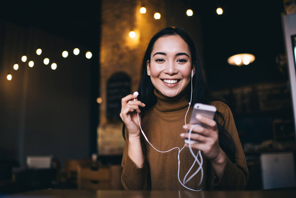 Junge lächelnde Frau in legerer Kleidung hört fröhlich Musik per Handy, während sie am Tisch im Café sitzt und in die Kamera schaut - Foto, Bild