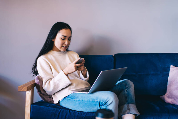 Side view of young positive woman in casual sweater and jeans browsing smartphone while working on laptop sitting on comfortable sofa - Foto, Imagem