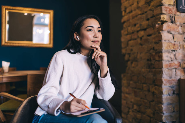 Concentrated Asian female writer in casual wear using true wireless earphones and taking notes in clipboard with sheet of paper while sitting in armchair in cafe and looking away - Фото, зображення