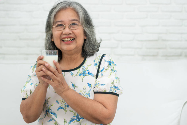 Asiática Senior mujer sosteniendo leche de vidrio mientras se relaja en un sofá sala de estar para el bienestar de jubilación. Anciana bebiendo un vaso de leche para mantener su bienestar. Concepto de bienestar y salud. - Foto, imagen