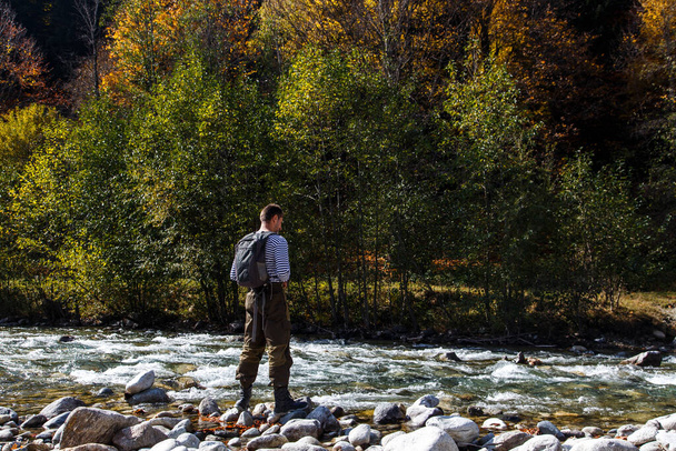 Człowiek z plecakiem stoi na skalistym brzegu górskiej rzeki plecami do kamery. Patrzy na przeciwny brzeg pokryty drzewami. - Zdjęcie, obraz