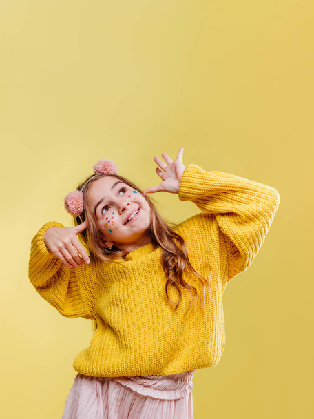 Funny teenage girl with stickers on her face wearing pink fluffy hair hood and yellow sweater having fun time on yellow background with blank space for text. - Zdjęcie, obraz