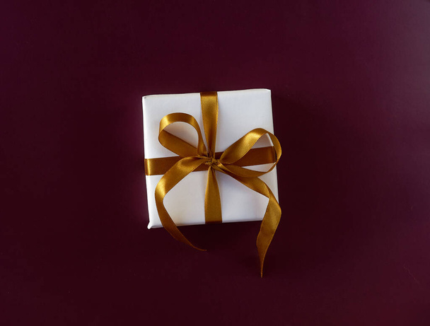 Cadeau doos verpakt in wit papier met een gouden strik op feestelijke violette achtergrond - Foto, afbeelding