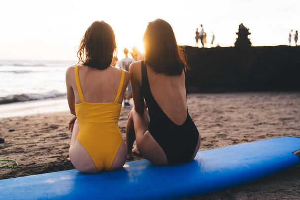 Back view of anonymous girlfriends resting on blue surfboard and observing sunny sky near wavy tranquil ocean on sandy beach - Photo, image