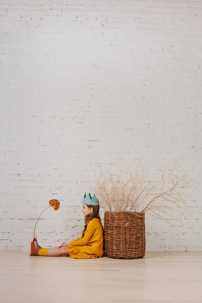 Bella bambina indossa corona seduta sul pavimento guardando fiori secchi ortensia. Vestito stile boho per bambini. Copia spazio per testo. - Foto, immagini