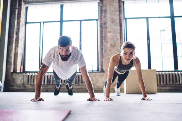 Determined male and female couple in love training together in modern gym keeping healthy lifestyle, handsome 20s bearded man trainer having workout with woman in active wear standing in plank - Fotó, kép
