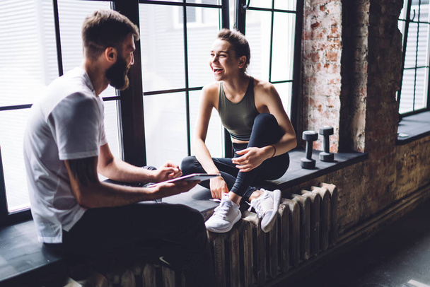 Cheerful young female in sports top and leggings having conversation with fit male holding tablet resting on industrial styled windowsill - Фото, изображение