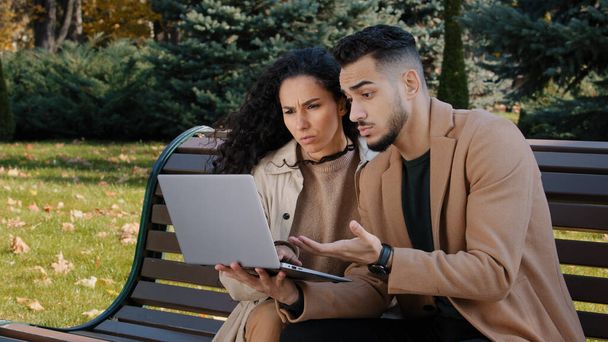 Worried hispanic young couple with laptop sit on bench in autumn park unsuccessful online shopping broken device serious frustrated business partners man and woman outdoors poor internet connection - Fotó, kép