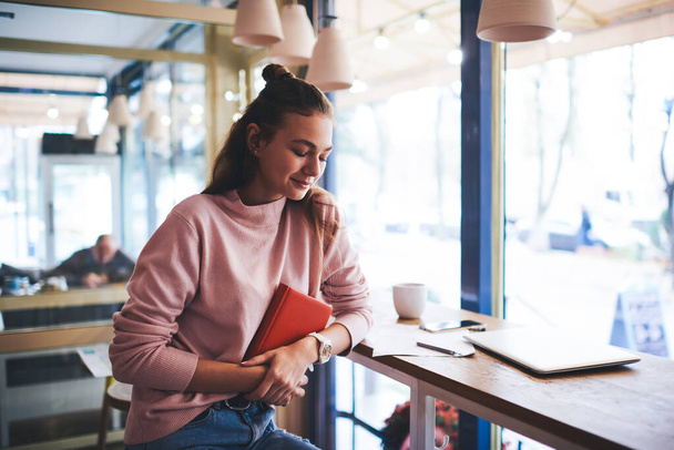 Positive female in casual outfit with long hair looking down sitting at table with book and cup of coffee having rest in cafe - 写真・画像