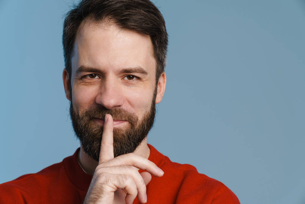 Brunette white man with beard smiling and showing silence gesture isolated over blue background - Фото, изображение
